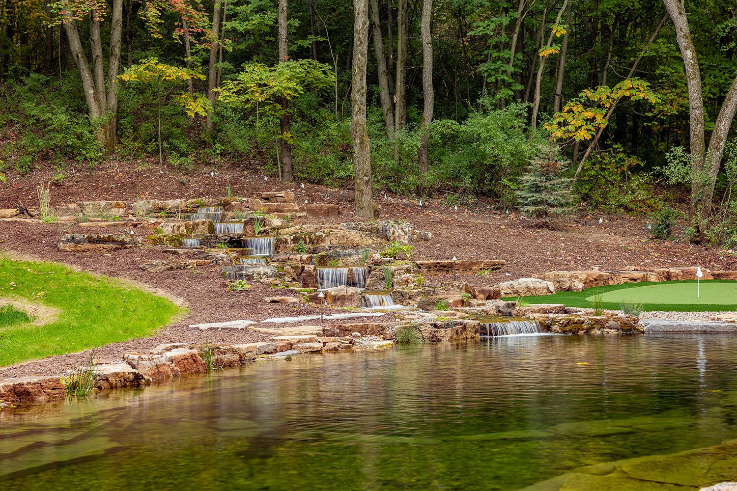 Backyard pond with multi-layer waterfall. There are also trees and putting green near the area.
