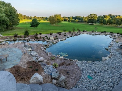 In the photo is a swim pound surrounded by rocks, green grass, and some trees. There's also a seating area on the side.